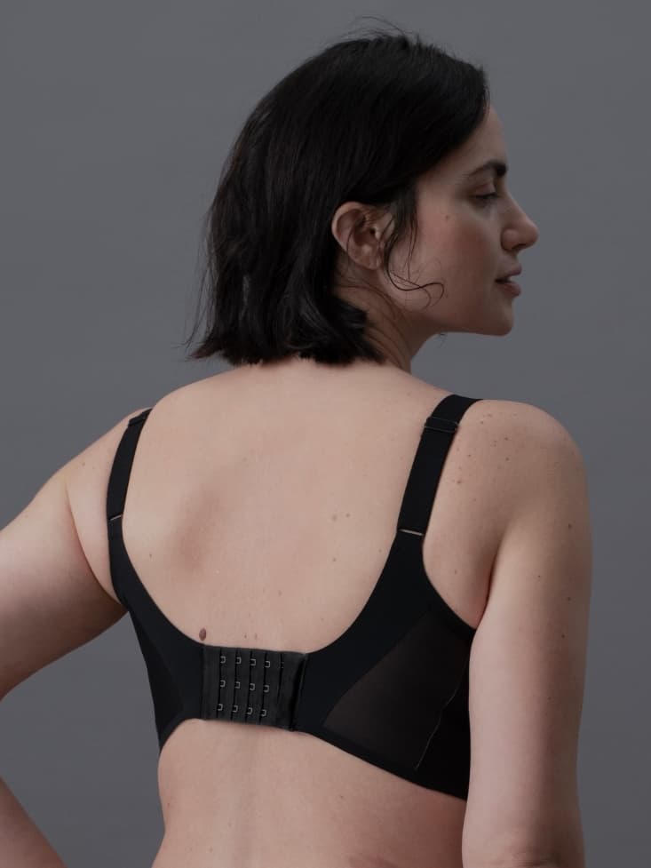 Woman with short dark hair, wearing a black bra with adjustable straps, seen from the back against a gray background.
