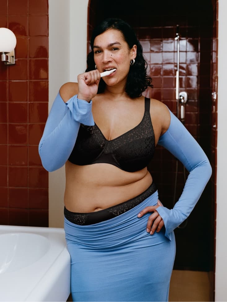 Person in a bathroom brushing their teeth, wearing a black bra and a blue outfit. The bathroom has maroon tiles and a sink.