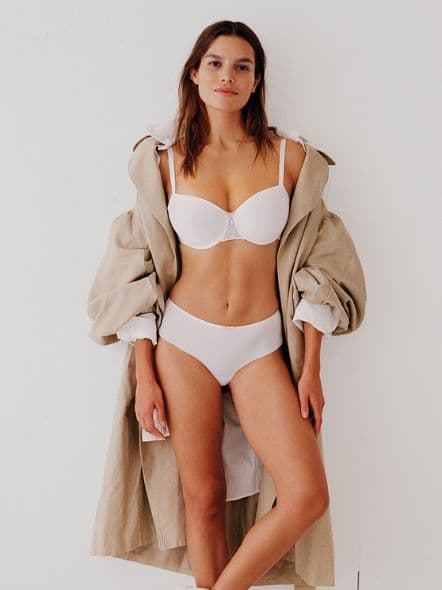 Woman in white lingerie and beige coat stands against a plain white background, smiling confidently.